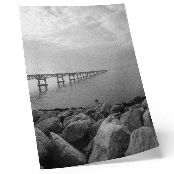 Wandposter vreedzaam landschap met een brug en een rotsachtig strand onder een grijze lucht